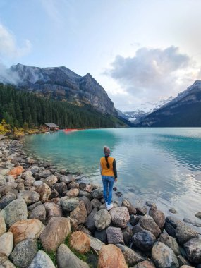 Kayalık sahilde duran bir figür, şafak vakti heybetli dağların çerçevelediği dingin sulara ve manzarayı aydınlatan sabahın yumuşak ışığına bakıyor. Louise Gölü Kanada