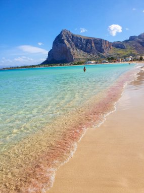 Turkuaz dalgalar, sığ sularda tekil bir figürün yürüdüğü kumlu bir kıyıya nazikçe yaklaşır. Arka planda çarpıcı bir dağ beliriyor, San Vito Lo Capo Sicilya