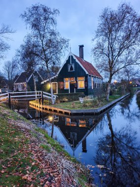 Resimli bir Hollanda evi durgun bir kanalda duruyor, ışıkları alacakaranlık gibi sıcacık parlıyor. Sonbahar yaprakları bankaları süslüyor, Zaanse Schans Hollanda