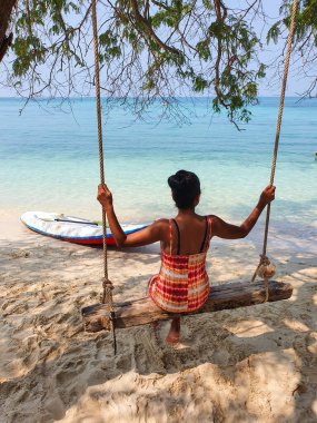 Kırsal bir salıncakta huzurlu bir anın tadını çıkaran bir kadın, sakin turkuaz sulara bakarken kumlu sahilde yumuşak güneş ışığı altında bir raket dinleniyor. Koh Kham Adası Chonburi Samaesan Tayland