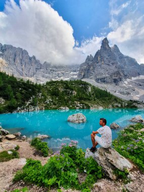 Nestled amidst towering peaks and lush greenery, a person gazes thoughtfully at the vibrant turquoise waters reflecting the blue sky, Lago Di Sorapis Dolomites Italy clipart