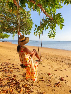 Renkli elbiseli genç bir kadın, sakin bir sahile bakan bir ağacın altında sallanarak huzurlu bir anın tadını çıkarıyor. Batan güneş, Krabi Tayland 'ın sıcak kum ve sakin sularına altın renkleri döküyor.