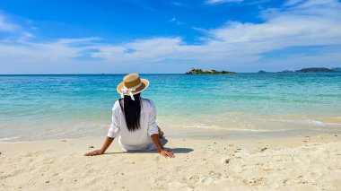 Yumuşak kumda oturup, Samae San Adası 'nın turkuaz sularına bakan, Tayland' da parlak mavi bir gökyüzünün altında, hafif dalgalar ve uzak adalarla çevrili, sakin bir an gözler önüne seriliyor.