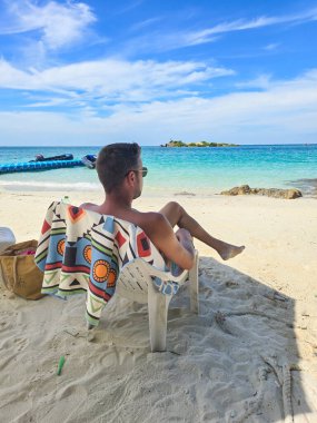 Samae San Adası 'nın huzurlu güzelliğinin tadını çıkaran bir gezgin, plastik bir sandalyede güneşlenerek oturur. Turkuaz sular ve yumuşak kum mükemmel bir tropikal geri çekilme atmosferi yaratır..