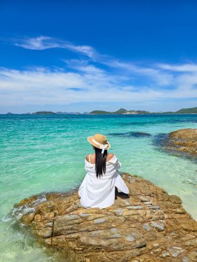 Enjoying the tranquil beauty of Samae San Island, a woman sits on a rock, gazing at the vibrant turquoise waters under a clear blue sky, Asian woman with hat relaxing on the beach in Thailand clipart