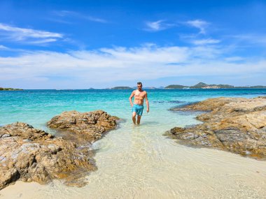 A traveler enjoys the serene beauty of Samae San Island, wading through shallow waters surrounded by stunning rock formations. Man in swim shorts on an Island near Pattaya Thailand clipart