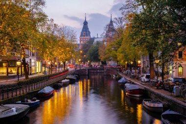 Alacakaranlık Amsterdam 'a çöktüğünde, sonbahar yapraklarının altın renklerini yansıtan kanallar parıldıyor. Samimi tekneler suyun kenarında, yoldan geçenleri huzur veren güzelliği keşfetmeye davet ediyor..