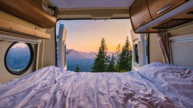 Bir karavanın içinde, güneş doğarken sıcak yatak örtüsü dışarıdaki nefes kesen dağları rahatlatıyor. Yemyeşil ağaçlar manzarayı çerçeveler, sakin Avusturya dağ manzarasında maceraya davetiye çıkarır..