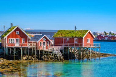 Ayaklıklardaki üç kırmızı kulübe Norveç fiyortlarında kayalık bir çıkıntının üzerinde duruyor ve turkuaz su açık mavi gökyüzünü yansıtıyor. Reine, Lofoten Adaları, Norveç