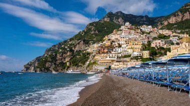 Amalfi kıyısında, Positano 'nun pitoresk kasabası kayalıklardan dökülen renkli binalar, kumlu sahil sandalyeleri mavi bir gökyüzü ve yumuşak dalgaların altında kıyıya dizilmiş..