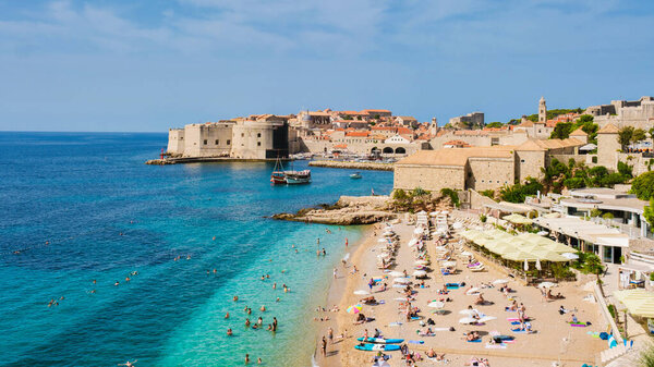 Посетители греются под теплым солнцем на песчаном пляже Дубровника, наслаждаясь кристально чистой водой и яркой исторической архитектурой вдоль берега Хорватии
