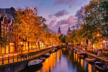 Alacakaranlık Amsterdam 'a inerken büyüleyici kanallar sonbahar ağaçlarının altın tonlarını yansıtıyor. Tekneler huzur içinde yüzerken dükkanlar sıcacık parlıyor. Amsterdam 'da Noel' de sokak lambaları yanıyor.