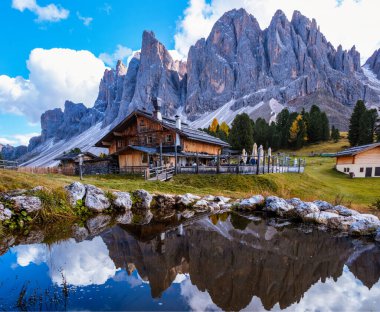 Çarpıcı Dolomitler, arka plandaki heybetli dağ sıralarını yansıtan sakin bir göletin yanında duran tuhaf ahşap kulübe. Geisleralm Dolomites Val Di Funes İtalya 'da Adolf Munkel Trail Autumn