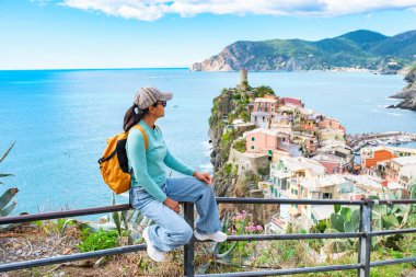 Bir kadın gezgin, Cinque Terre 'nin çarpıcı sahil şeridine bakarak bir korkulukta oturur. Uçurumlara tünemiş canlı evler ve ışıl ışıl deniz manzaralı bir zemin oluşturuyor, Vernazza İtalya