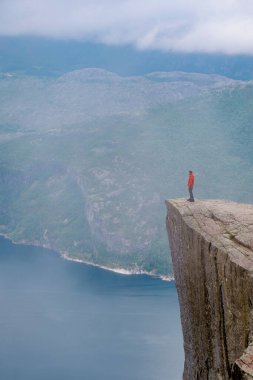 Bir yürüyüşçü, kolları açık bir uçurumun kenarında durur ve sisle kaplanmış Norveç fiyortlarının nefes kesici manzarasını seyreder. Önceki bölümlerde, Norveç 'te yazın