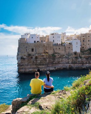 Polignano a Mare Italy, A couple sits together on the rocky cliff, gazing at the picturesque white buildings of Puglia, bathed in sunlight. a diverse couple of men and women on vacation clipart