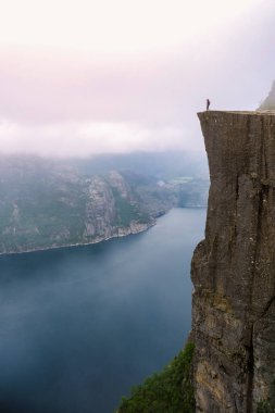 Preikestolen 'in kenarında yalnız bir birey duruyor. Norveç' te fiyorda tepeden bakan bir uçurum. Gökyüzü sisli ve dramatik. Önceki bölümlerde Norveç, stres, hayal kırıklığı ve tükenmişlik ya da başarı kavramı