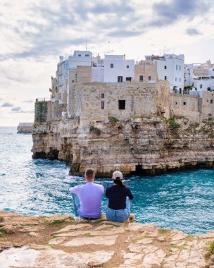 A couple sits peacefully on rocky terrain, gazing at the turquoise waters and ancient cliffs of Puglia, Italy. The sun begins to set on the charming coastal village. Polignano a Mare Italy clipart