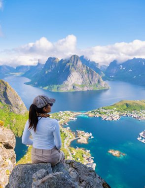 Norveç Lofoten Adaları 'nın büyüleyici fiyortlarına ve dağlarına bakan bir kadın uçurumda oturuyor. Temiz mavi su ve yemyeşil tepelerle manzara nefes kesici. Reinebringen Norveç