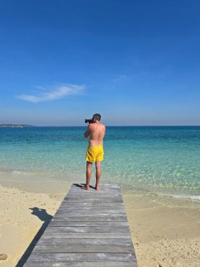 Tahta bir iskelede duran yalnız bir figür Koh Munnork Adası 'nın büyüleyici turkuaz sularına hayran. Altın kum ve parlak mavi gökyüzüyle çevrili bu an huzur dolu adanın mutluluğunu temsil ediyor..