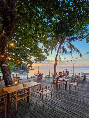 Guests gather on a wooden deck under swaying palms, savoring delightful meals as the sun sets over the sparkling waters of Koh Munnork Island, creating a magical atmosphere in Thailand. clipart