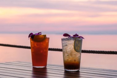 Güneş Koh Munnork Adası Tayland 'ın sakin sularının üzerinde batarken, renkli çiçeklerle süslenmiş ahşap bir masada iki canlı kokteyl oturmaktadır. Rayong 'da mükemmel bir akşam..