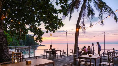Sallanan palmiye ağaçları ve tel lambaları arasında, restoranlar Koh Munnork Adası 'nda sakin bir akşam yemeğinin tadını çıkarıyorlar. Parlak günbatımı, turuncu ve pembe tonlarıyla gökyüzünü boyar ve dingin bir atmosfer yaratır..