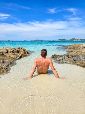 Tayland 'ın Samae San Adası' nın dingin güzelliğinin tadını çıkaran bir adam, yumuşak kumda otururken turkuaz dalgalar parlak güneşin altındaki kayalara hafifçe çarpıyor..