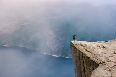 Bir yürüyüşçü, kolları açık bir uçurumun kenarında durur ve sisle kaplanmış Norveç fiyortlarının nefes kesici manzarasını seyreder. Önceki bölümlerde, Norveç 'te yazın