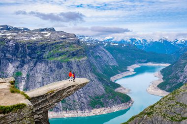 İki yürüyüşçü, Norveç 'in Trolltunga kentindeki dramatik fiyort ve dağ manzarasına bakan bir uçurumun kenarında duruyor. Dağlarda yürüyüş yapan bir çift adam ve kadın.