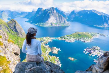 Asyalı bir kadın, dağları, berrak suları ve aşağıdaki küçük bir kasabayla Norveç fiyortlarını gören kayalık bir uçurumda oturuyor. Reinebringen, Lofoten, Norveç