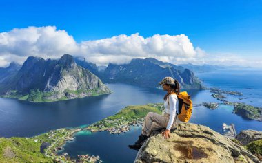 Asyalı bir kadın kayalık bir çıkıntıya tünemiş, Lofoten Adaları 'nın büyüleyici fiyortlarına ve tepelerine bakıyor güneş doğarken, nefes kesici manzarayı aydınlatıyor. Reinebringen Lofoten Norveç