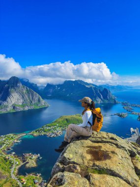 Bir yürüyüşçü, Lofoten Adaları 'nın görkemli dağları ve canlı mavi sularla çevrili nefes kesici manzaralarına hayran bir şekilde bir kayanın üzerinde oturur. Reinebringen Lofoten Norveç. Sırt çantalı Asyalı kadınlar
