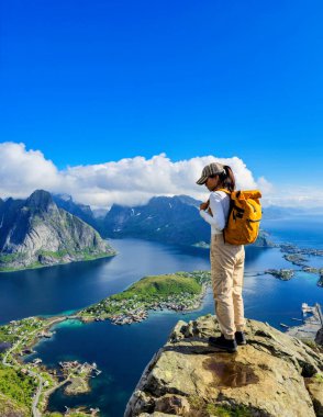 Yalnız bir yürüyüşçü engebeli bir uçurumun tepesinde duruyor, açık mavi bir gökyüzünün altında Norveç Lofoten Adaları 'nın nefes kesici manzaralarına dalmış. Asyalı kadınlar Lofoten 'i ziyaret ediyor
