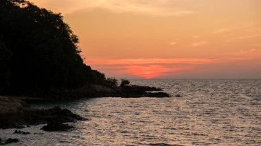 Gün ışığı söndükçe, gün batımının canlı renkleri Koh Munnork Adası 'nın üzerindeki gökyüzünü boyuyor. Tayland 'da akşam karanlığında yapraklar parıldarken kayalık kıyı şeridine hafif dalgalar çarpar..