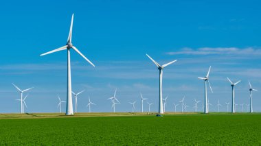 Uzun rüzgar türbinleri açık mavi gökyüzüne karşı zarifçe döner, yeşil tarlalara tepeden bakarken yenilenebilir enerjiyi kullanırlar. Yel değirmeni parkı Hollanda 'da yeşil enerji üretiyor.