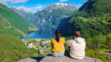 Sersemletici bir Norveç fiyorduna bakan kayalık bir çıkıntıda oturan iki kişi, yemyeşil dağlarla ve açık mavi gökyüzüyle çevrili. Geiranger Fjord Norveç. Birçok erkek ve kadın tatilde.