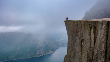 Bir çift, bir uçurumun kenarında durur ve sisle kaplanmış Norveç fiyortlarının nefes kesici manzarasını seyrederler. Preikestolen, Norveç Yazın. Erkekler ve kadınlar bir dağın tepesinde