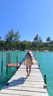 Bir kadın, yemyeşil sularla çevrili kristal berrak suların üzerinde uzanan ahşap bir iskelede yalınayak yürüyor. Gökyüzü, Koh Kood Tayland 'ın dingin güzelliğine ekleniyor.