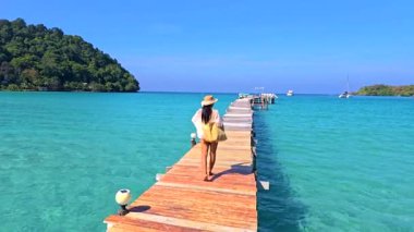 Koh Kood 'un güzelliğinin tadını çıkarın. Bir kadın, turkuaz sulara uzanan ahşap bir iskele boyunca geziniyor. Bereketli yeşilliğin ve Tayland 'daki parlak gökyüzünün yakınındaki huzuru hisset.