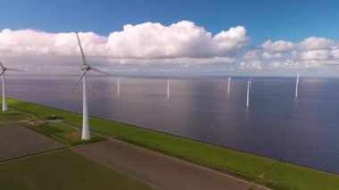 Hollanda kıyıları boyunca uzanan yenilenebilir enerji türbinleri mavi gökyüzü ve kabarık bulutlara karşı sürdürülebilir enerji sağlıyor. Okyanustaki yel değirmeni parkı, yel değirmeni türbinlerinin insansız hava aracı görüntüsü. 