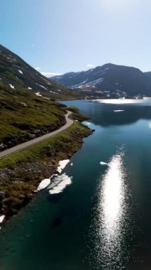 Norveç 'te nefes kesici manzaraları keşfetmek için sakin, ışıl ışıl bir göl boyunca uzanan pitoresk bir yol kıvrımları keşfedin. Karla kaplı dağlar sakin suları yansıtıyor, Langvatnet Geiranger Norveç