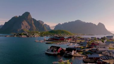 Norveç 'in pitoresk bir kıyı köyü, canlı kırmızı kulübeler, suda hafifçe sallanan kayıklar ve gün batımının sıcak parıltısıyla aydınlanan görkemli dağlarla gözler önüne seriliyor. Reine Köyü Lofoten