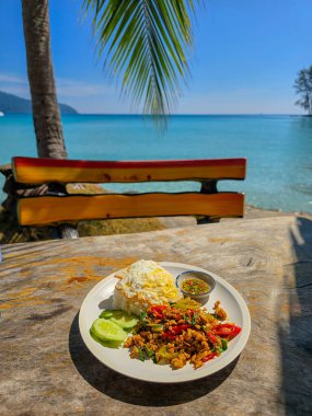 A vibrant plate of Thai cuisine Phat kaphrao rests on a rustic wooden table, with glistening turquoise waters and palm trees in the background. The perfect spot for an unforgettable dining experience. clipart