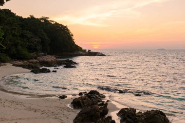 Güneş Koh Munnork Adası 'nda batarken dalgalar yavaşça kayalık sahile vurur. Gökyüzünü turuncu ve mor renklerle boyarlar..