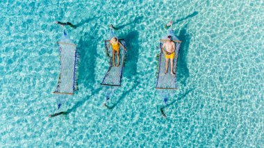 Two individuals lounge on floating mats in crystal-clear turquoise waters of Koh Kood. couple of men and a woman relaxing in a hammock above the ocean clipart