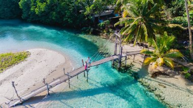 A couple strolls along a wooden bridge connecting lush greenery and pristine waters in Koh Kood, Thailand. The warm hues of dawn create a peaceful ambiance, inviting exploration and tranquility. clipart