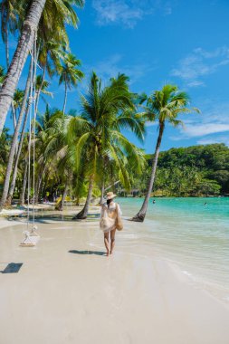 Koh Kood 'un yumuşak kumlu plajında yürüyen bir kadın, sakin bir atmosfer kıyıda hafif dalgalar tur atarken etrafını sarıyor. Ilık güneşte sallanan gür palmiye ağaçları resim gibi bir kaçış yaratıyor..