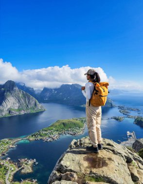 Bir yürüyüşçü engebeli bir uçurumun tepesinde duruyor, güneş batarken Norveç 'in büyüleyici fiyortlarına bakıyor, dingin suları aydınlatıyor. Reinebringen Lofoten Norveç, Lofoten 'de sırt çantalı Asyalı kadınlar yürüyüş yapıyor.