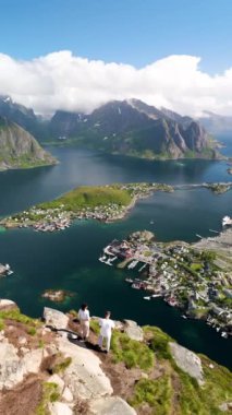 Norveç 'teki sakin bir fiyort köyünün üzerinde görkemli dağlar yükseliyor. İki yürüyüşçü kayalık bir çıkıntıda duruyor, yemyeşil yamaçların panoramik manzarasını izliyorlar, Reinebringen yürüyüşü Lofoten Norveç